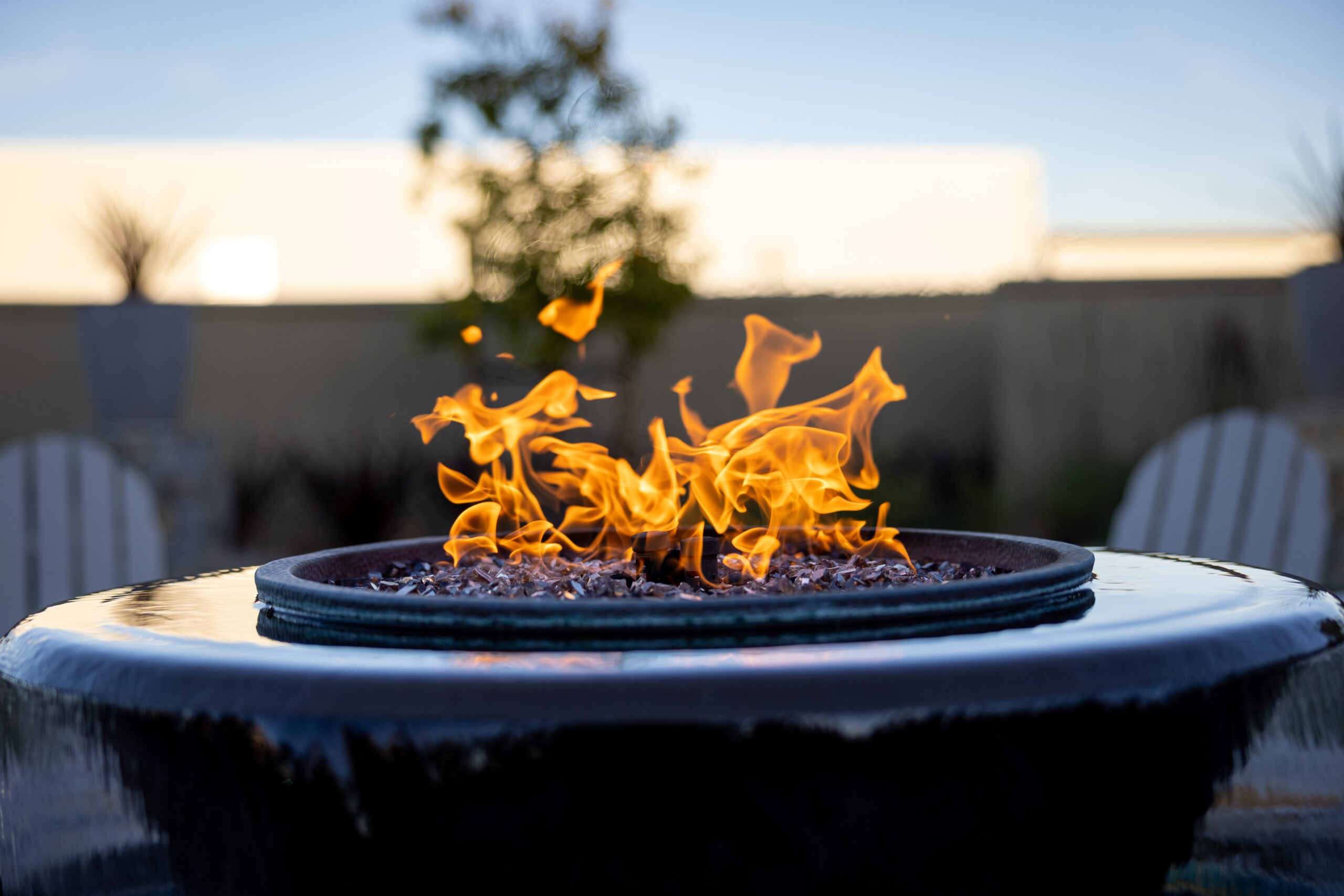 A vibrant fire burns in the centre of a round gas firepit table with a blurred background of a fence and plants during twilight, highlighting the warm flames against the cool dusk sky.
