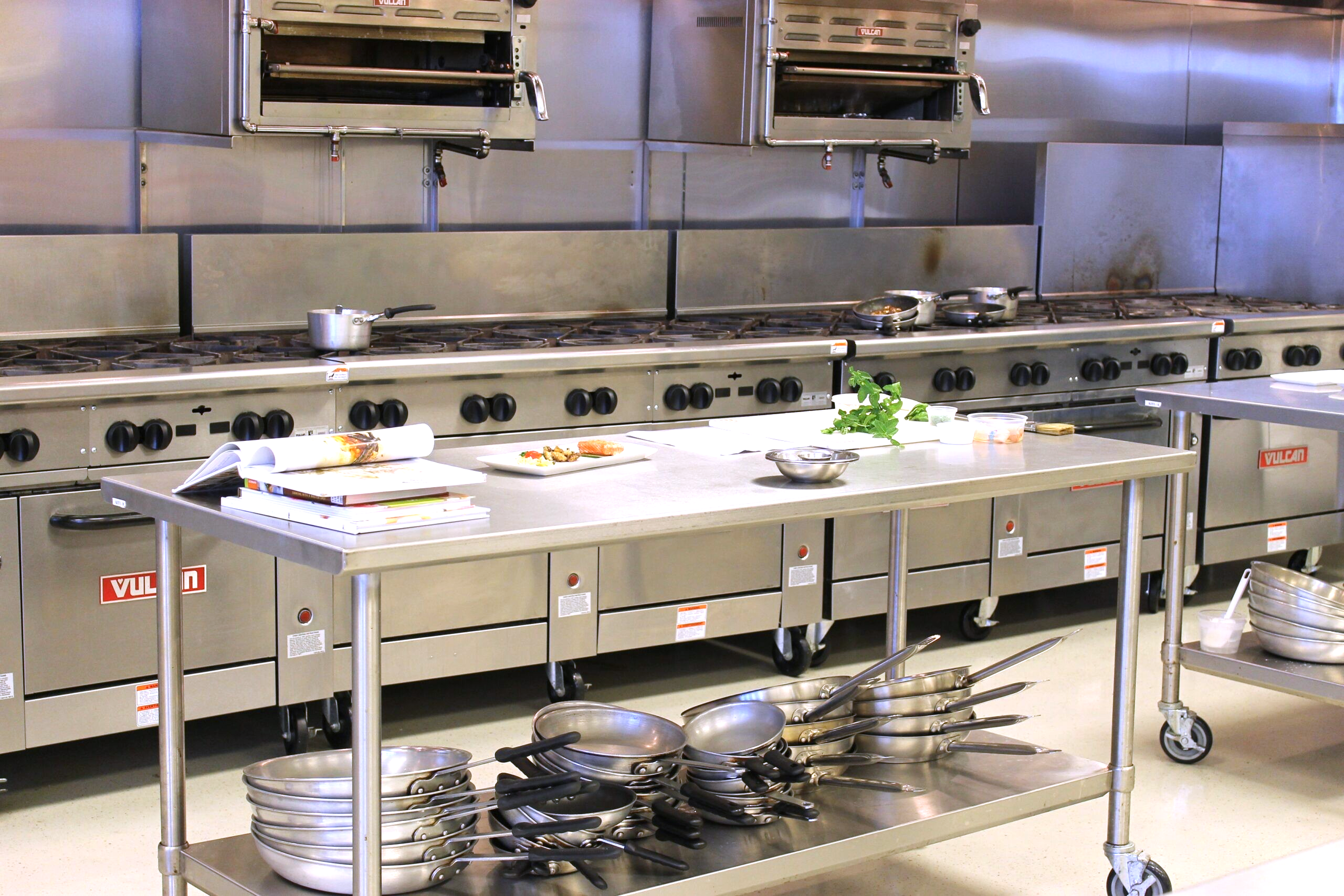 A professional kitchen with stainless steel equipment. There are multiple stovetops and ovens along the back wall, with a large stainless steel preparation table in the center holding various cooking utensils, a stack of plates, and some scattered green vegetables. To the right, there is a mobile stainless steel cart with more plates on it. The kitchen appears clean and well-organized, ready for meal preparation.