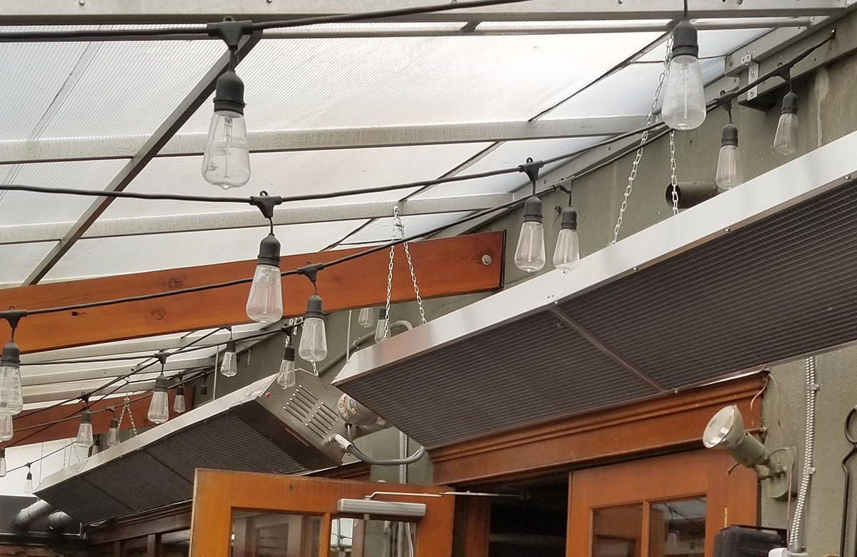 The image displays a series of tube heaters installed overhead. Each heater is encased within an emitter guard, which appears to be a metal grid or mesh surrounding the tube to prevent direct contact with the heating element. The heaters are suspended from a structure with chains.