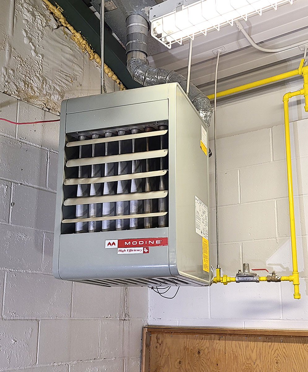 A Modine gas-fired unit heater is mounted on a concrete wall in a utility room. The heater features horizontal louvers and is connected to an exhaust pipe extending into the ceiling. To the right, a yellow gas pipe with a red valve runs vertically and bends into the unit. The background shows exposed brickwork and conduits, indicating an industrial or commercial setting.