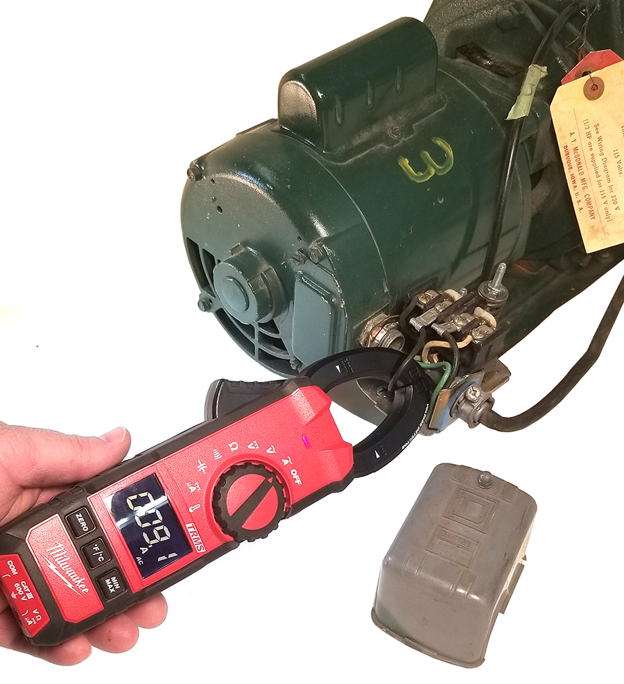A digital clamp meter displaying a reading of 0.09 is clamped around a single electrical wire connected to a large green industrial motor. The motor has a yellow tag attached, and there’s an unattached metal cover plate resting to the right side. A hand is holding the clamp meter, indicating the measurement of current flow or electrical continuity in the setup.