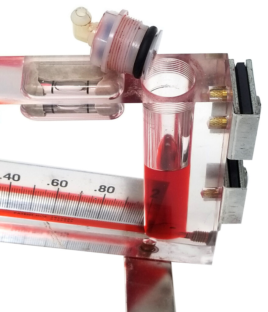 Close-up image of a Dwyer inclined manometer, focusing on the top section where the red gauge oil is filled. The image shows the fluid level and the calibration scale in inches of water, with the top cap and connector piece removed, revealing the threaded opening.