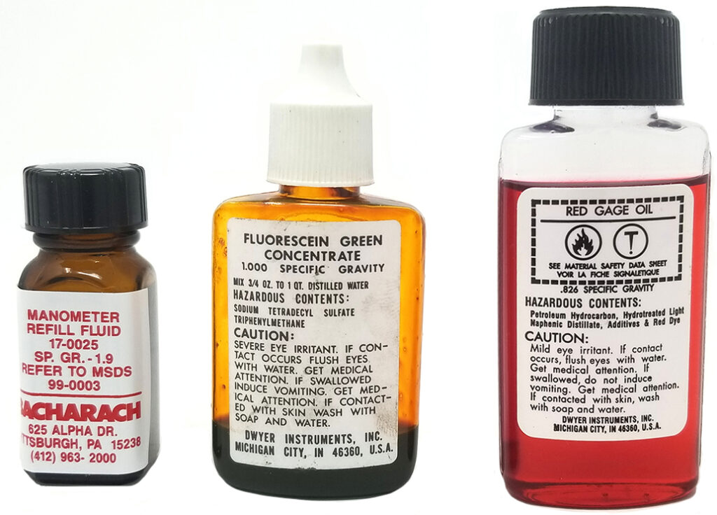 Image showing three bottles of different fluids: Manometer Refill Fluid, Fluorescein Green Concentrate, and Red Gage Oil. Each bottle has a label indicating specific gravity and hazard warnings, with the manometer refill fluid having a specific gravity of 1.9, the fluorescein green concentrate with a specific gravity of 1.000, and the red gage oil with a specific gravity of .826.