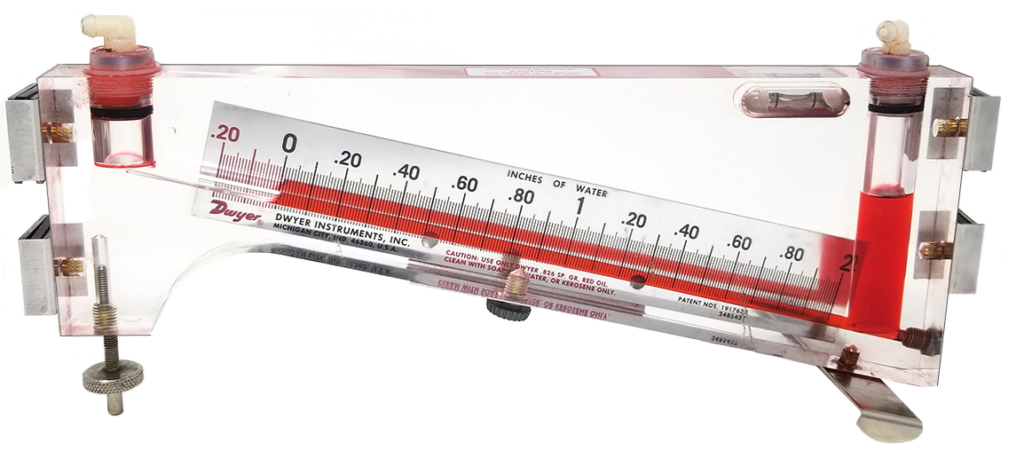 Image of a Dwyer Instruments inclined manometer, calibrated in inches of water. The manometer is filled with red gauge oil, with the scale displaying pressure measurements ranging from -20 to +20 inches of water.